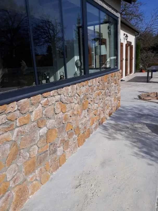 Grande baie vitrée à châssis aluminium fins posée sur un soubassement en parement de pierre, vue depuis la terrasse d'une maison de plain-pied